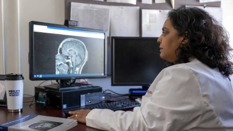 Dr. Bavishi pictured wearing a white lab coat and looking at a compute with a picture of a brain scan